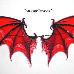 May include: A pair of red and black decorative wings with a glittery finish. The wings have a lace-like pattern and are connected by a black chain. The text "*indigo* moon*" is above the wings.