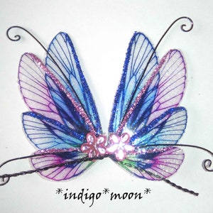 May include: A pair of fairy wings in shades of blue, purple, and pink, with glitter accents. The wings have a delicate, translucent appearance and are adorned with small pink flower embellishments. The text "indigo*moon*" is at the bottom.