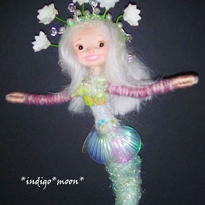 Op de afbeelding: Handgemaakte zeemeerminnenpop met wit haar en een lachend gezicht. De pop heeft een glinsterende, iriserende staart en een kroon van witte bloemen. De armen zijn gewikkeld in roze garen. De tekst "*indigo*moon*" is zichtbaar.