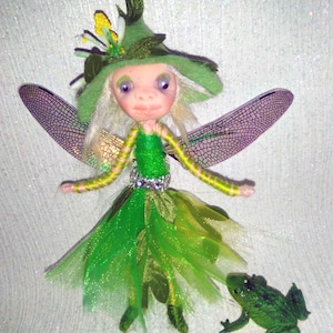 Puede incluir: Muñeca de hada hecha a mano con vestido verde, sombrero y alas de libélula. Una pequeña figura de rana está cerca. El texto "*indigo* moon*" está escrito en la parte superior de la imagen. Diseño caprichoso.