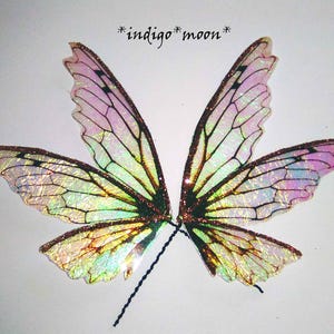 Op de afbeelding: Iriserende feeënvleugels met een glitterafwerking. De vleugels hebben een kleurengradient, waaronder roze, groen en goud, met zwarte aderachtige details. De tekst "*indigo*moon*" staat boven de vleugels.