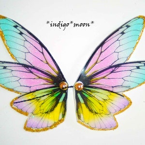 Op de afbeelding: Een paar iriserende feeënvleugels met een kleurengradatie van blauw, roze, geel en groen. De vleugels hebben een gouden glitterrand en zijn verbonden door twee goudkleurige sluitingen. De tekst "*indigo* moon*" staat boven de vleugels.