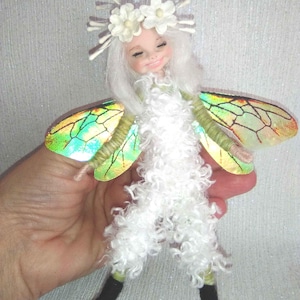 Puede incluir: Una muñeca de hada hecha a mano con un cuerpo blanco rizado, una corona floral y alas iridiscentes. La muñeca tiene una cara sonriente y lleva botas negras. El texto "*indigo*moon*" está en la esquina superior izquierda.