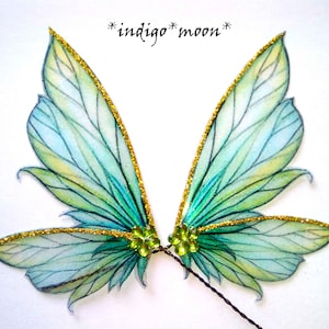 May include: A pair of fairy wings with a delicate design. The wings feature a gradient of blue and green hues, outlined with gold glitter. The text "*indigo* moon*" is above the wings. The wings are connected by a small green gem.