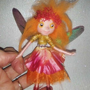 May include: A handmade fairy doll with orange hair and a red floral crown. The doll wears a gold top and a layered pink and orange skirt. It has dragonfly-like wings and is held in a hand.