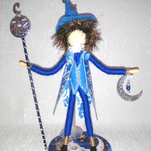 May include: A handcrafted wizard doll in blue attire with white floral patterns, holding a silver staff and a crescent moon. The doll stands on a circular base with a moon and star design. The text "*indigo* moon*" is visible.