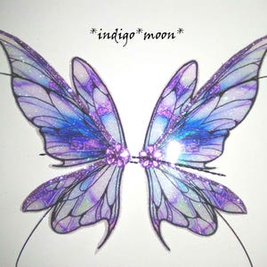 May include: A pair of fairy wings with a shimmering design. The wings feature a gradient of purple and blue hues, with glitter accents. The text "*indigo moon*" is written above the wings. The wings are connected by a central jewel.