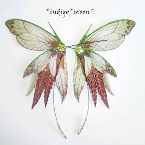 Puede incluir: Un par de alas de hada con un diseño delicado. Las alas presentan una combinación de elementos transparentes, brillantes y marrones, con acentos verdes. El texto "*indigo*moon*" es visible en la parte superior. Las alas están diseñadas para ser usadas.