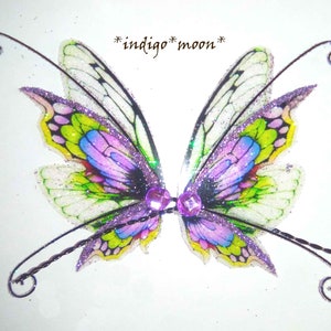 May include: A pair of iridescent fairy wings with purple, green and white glitter. The wings are attached to a wire that forms a loop at each end.