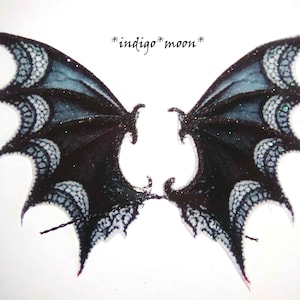 May include: A pair of black and white lace wings with a glittery finish. The wings are shaped like a dragon's wings and have a delicate, intricate design. The text "*indigo*moon*" is written in cursive at the top of the image.