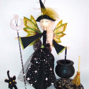 Op de afbeelding: Een handgemaakte feeënheks pop met een zwarte jurk versierd met sterren en manen. De pop heeft blond haar, groene en gele vleugels en een zwarte hoed met veren. Accessoires zijn een toverstaf, ketel, bezem en een zwarte kat.