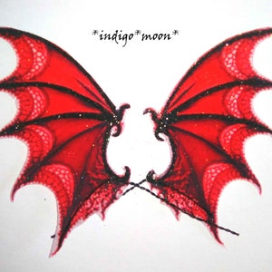 May include: A pair of red, translucent fairy wings with black accents and glitter. The wings are connected by a black string and feature the text "*indigo *moon*". The wings have a detailed, veined pattern.