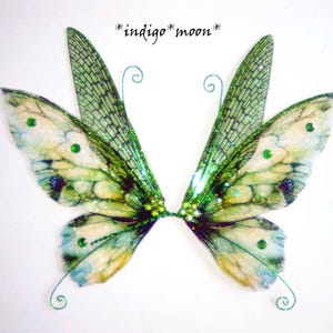 May include: A pair of fairy wings with a delicate design. The wings feature a gradient of colors, including green, white, and blue, with glitter accents. The text "*indigo* moon*" is written in black script above the wings. The wings are adorned with green gems.