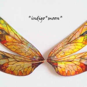 Puede incluir: Un par de alas de libélula iridiscentes con un diseño de vitral. Las alas son una mezcla de amarillo, naranja, rojo, azul y verde. Las alas están conectadas por un fino hilo dorado. El texto "*indigo*moon*" está escrito en cursiva negra sobre las alas.
