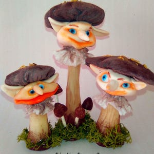 Puede incluir: Tres figuras de hongos caprichosas con caras de duendes, que llevan sombreros marrones y cuellos con volantes, se alzan sobre un lecho de musgo verde. Las caras están pintadas con ojos azules y acentos naranjas. El texto "indigo moon" está en la parte inferior.