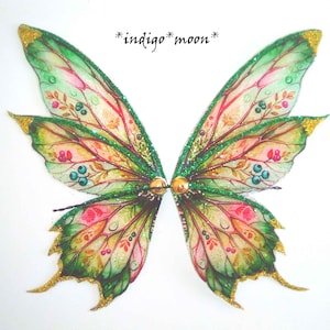 May include: A pair of fairy wings with a colourful design. The wings feature a green, pink, and yellow colour scheme with gold glitter accents. The text "*indigo* moon*" is visible in the upper right corner.