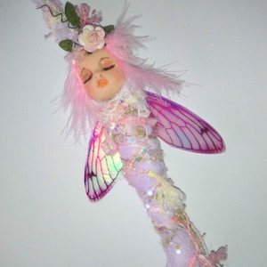May include: A whimsical fairy wand with a serene face, pink hair, and delicate wings. The wand is adorned with flowers, feathers, and sparkling embellishments. The text "*indigo* moon*" is at the top.