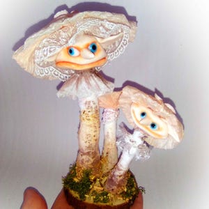 Peut inclure: Deux figurines de champignons fantaisistes avec des visages expressifs et des yeux bleus. Les champignons ont des chapeaux crème bordés de dentelle et reposent sur une base de mousse et de bois. Le texte "indigo moon" est en haut.