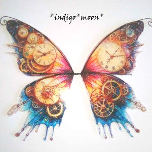 May include: A pair of steampunk-style fairy wings with a clockwork design. The wings feature a gradient of colors, including blue, pink, and orange, with intricate clock faces and gears. The text "*indigo* moon*" is visible.