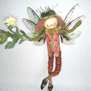 Op de afbeelding: Een grillige feeënpop met een groene kroon, bruin haar en een snor. De fee draagt een vest met patroon en oranje benen. Ze houdt een bloem vast en heeft delicate vleugels. De tekst "indigo*moon*" staat bovenaan.