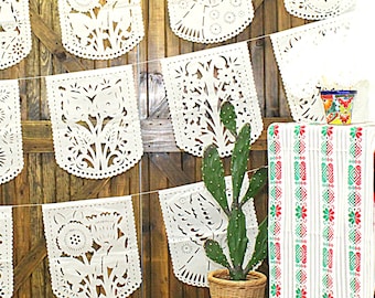May include: A string of white paper cutouts with intricate floral and bird designs hangs against a rustic wooden background. A small potted cactus and a colorful table runner with green and red stripes are in the foreground.