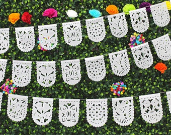 May include: A white paper garland with a scalloped edge and intricate cut-out designs. The garland is strung with white thread and features various patterns, including butterflies, flowers, and geometric shapes. The garland is hanging against a backdrop of green foliage.