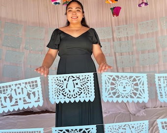 May include: A long dark green dress with short flutter sleeves and a V-neckline, positioned behind a string of light blue paper banners featuring intricate cut-out designs of birds, suns, and cacti, with colorful paper decorations hanging above.