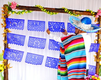 May include: A series of blue paper banners with intricate cut-out floral and geometric patterns hanging on a wooden frame. A person wears a blue and silver sombrero and a vibrant, multi-colored striped serape over a white long-sleeve shirt.