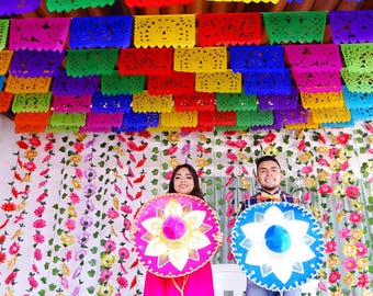 May include: Two people holding circular sombreros, one pink and one blue, against a backdrop of colorful cut-paper banners and hanging floral garlands. The sombreros feature intricate white floral patterns and bright, solid-colored crowns.
