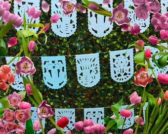May include: A string of blue paper banners with cut-out designs hangs against a backdrop of green foliage. Pink and red tulips are arranged around the banners, creating a festive and colorful scene.