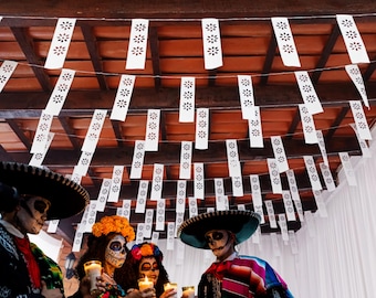 May include: A festive Day of the Dead scene with people in traditional attire and face paint, holding candles. Overhead, white paper banners with floral cutouts hang from a wooden structure, creating a celebratory atmosphere.