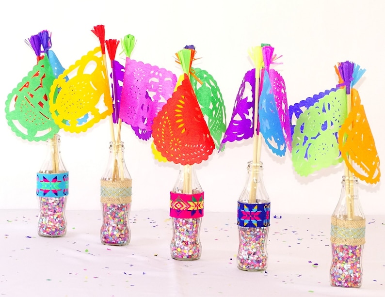 May include: Five glass bottles filled with colourful confetti and topped with colourful paper cutouts, resembling flags, with tassels. The bottles are decorated with colourful patterned bands.