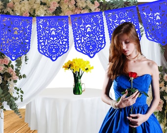 May include: Blue paper cutouts with intricate designs hang from a white sheer curtain. A woman in a blue dress holds a single red rose. A white table with a white tablecloth is in the background.
