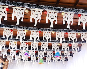 May include: A festive display of white paper cutouts with star-shaped centers, strung across a wooden ceiling. Red and gold ornaments are interspersed among the cutouts, with green and silver tinsel adding a touch of sparkle. Colorful paper bells hang below.