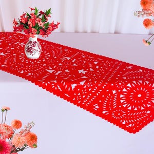 May include: A vibrant red table runner with intricate cut-out floral patterns. A white vase with floral designs holds red flowers, complementing the runner's color. The runner is placed on a white surface, with additional flowers in the background.
