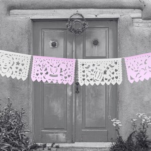 May include: A string of white and pink paper banners with cut-out designs hangs in front of a grey door. The banners are decorated with hearts, stars, and other shapes.