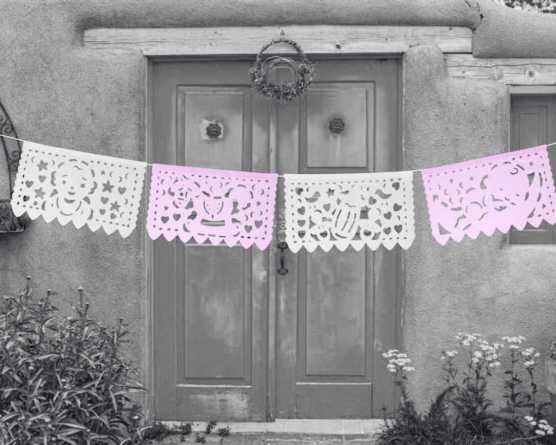 May include: A string of white and pink paper banners with cut-out designs hangs in front of a grey door. The banners are decorated with hearts, stars, and other shapes.