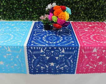 May include: Three colorful papel picado table runners in light blue, blue, and pink. Each runner features intricate cut-out designs of flowers, birds, and geometric patterns. A vibrant bouquet of flowers sits atop the blue runner, adding a festive touch.