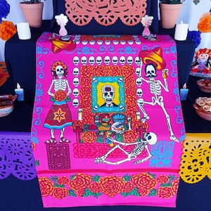 May include: A vibrant pink Day of the Dead altar cloth featuring skeleton figures, skulls, and floral patterns. The design includes a central framed skull portrait, surrounded by roses and decorative borders. The cloth is set on a dark blue surface.
