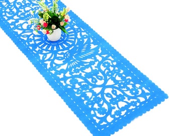 May include: A vibrant blue table runner with a detailed, openwork design featuring floral and bird motifs. The runner has a scalloped edge and is placed on a white surface. A small white vase with flowers sits on top.
