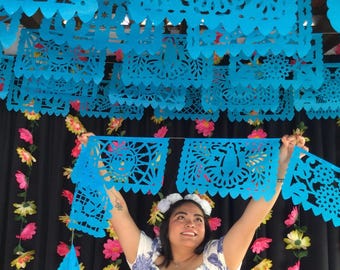 May include: A string of bright blue papel picado banners featuring intricate cut-out designs of suns and birds. The rectangular paper panels have scalloped edges and are suspended horizontally, showcasing detailed patterns created through precise paper cutting.