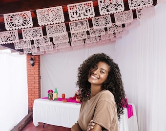 May include: A woman smiles in front of a table set for a celebration. Overhead, white paper banners with intricate cut-out designs hang from the ceiling. The table is covered with a white tablecloth and a pink runner, with a bottle and glasses.