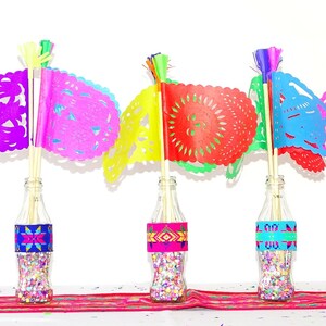 May include: Five glass bottles filled with confetti and decorated with colourful patterned paper bands. Each bottle has a colourful paper flag with a cut-out design attached to a wooden stick.