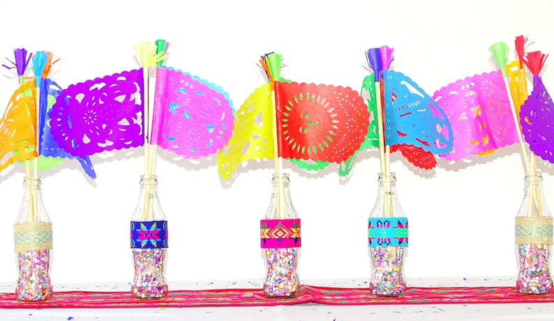 May include: Five glass bottles filled with confetti and decorated with colourful patterned paper bands. Each bottle has a colourful paper flag with a cut-out design attached to a wooden stick.