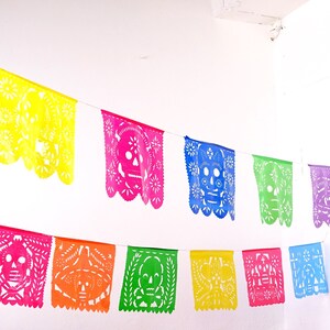 May include: A colorful paper banner with cut-out skull designs in various colors, including yellow, pink, blue, green, and orange. The banner is strung up against a white wall.
