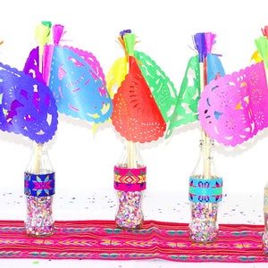 May include: Five glass bottles filled with colourful confetti and topped with colourful paper flags. The bottles are decorated with colourful patterned fabric bands. The bottles are arranged on a red and white striped table runner.