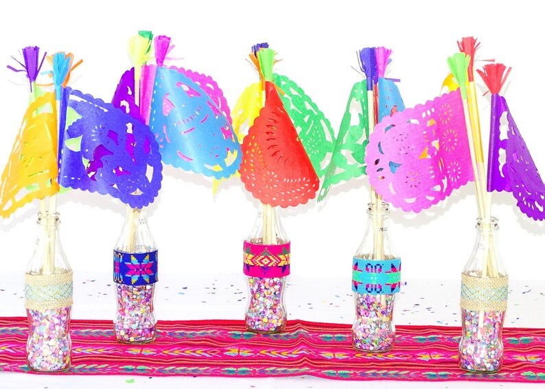 May include: Five glass bottles filled with colourful confetti and topped with colourful paper flags. The bottles are decorated with colourful patterned fabric bands. The bottles are arranged on a red and white striped table runner.