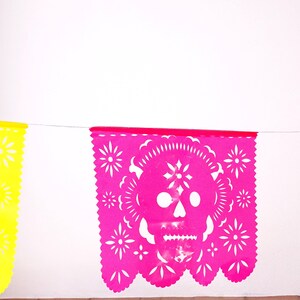 May include: A string of colorful paper banners with cut-out designs. The center banner features a pink skull with white details. The banners are yellow, pink, and blue.