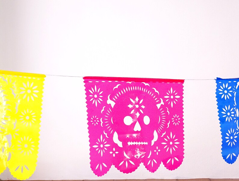 May include: A string of colorful paper banners with cut-out designs. The center banner features a pink skull with white details. The banners are yellow, pink, and blue.