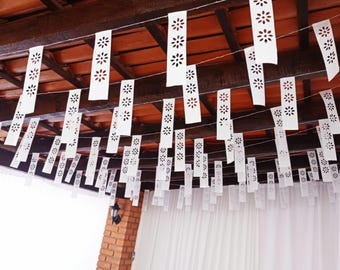 May include: White paper banners with floral cut-out designs are strung across a ceiling. The banners hang from a wooden beam ceiling with a red tile roof. The scene is lit by a wall-mounted light fixture on a brick pillar.
